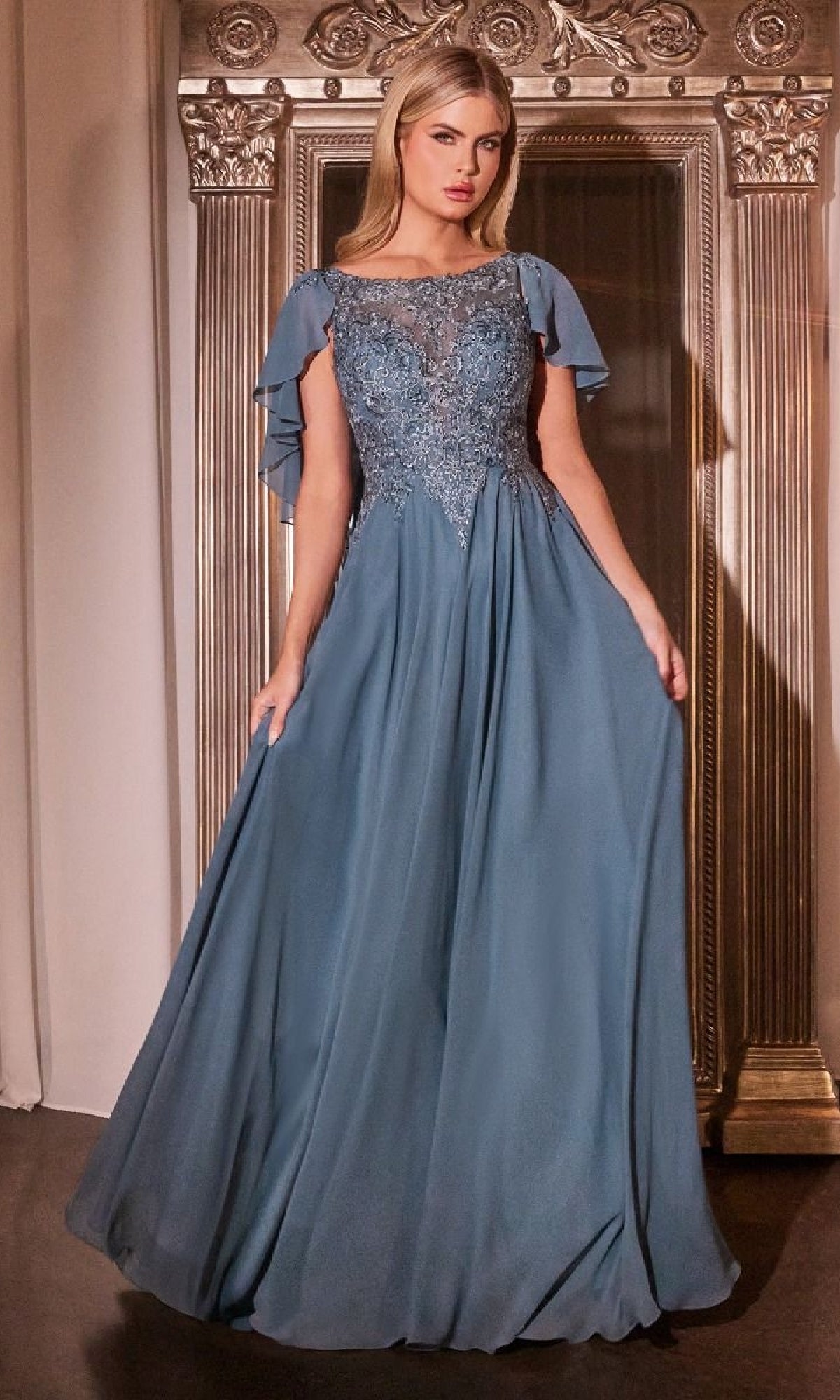 A woman stands before an ornate mirror wearing the Ladivine HT101, a floor-length blue chiffon A-line prom dress featuring lace bodice details and elegant cape sleeves.